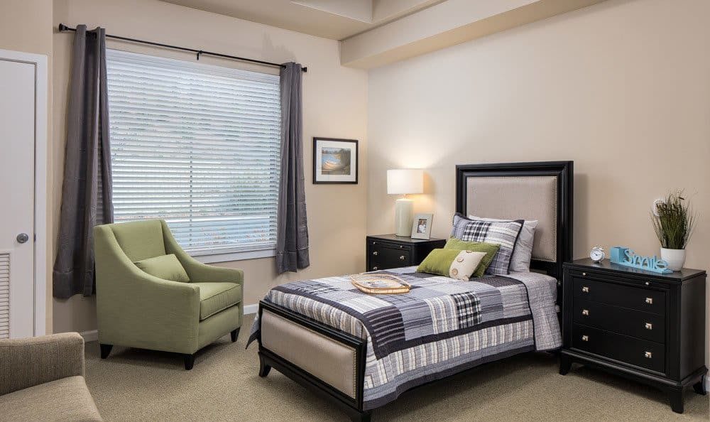 A cozy bedroom featuring a single bed with a patterned quilt, a green armchair, and soft lighting from a table lamp.
