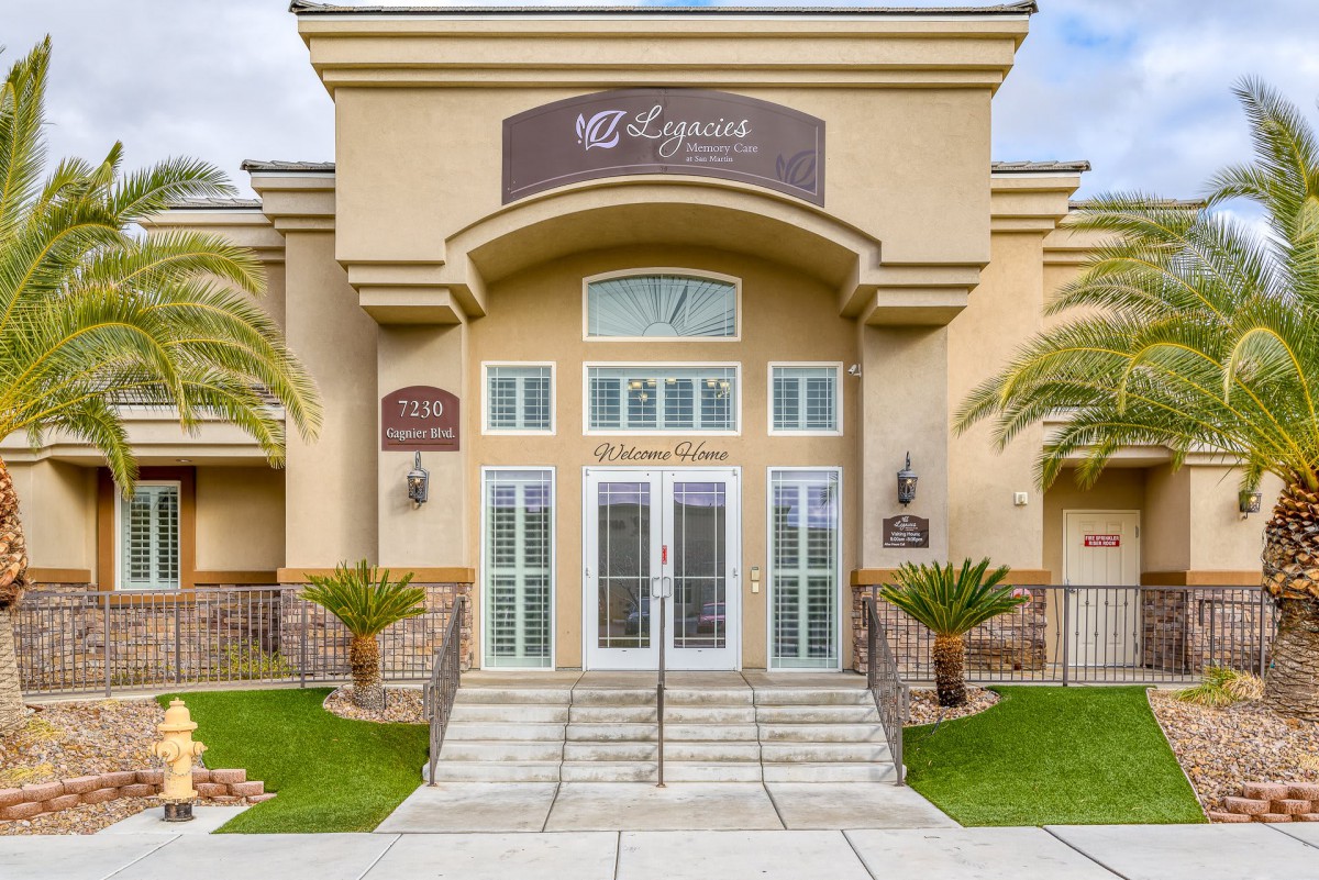 The front entrance of a beige building with glass doors and palm trees on either side.