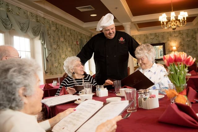 A chef interacts with elderly guests at a restaurant table, discussing the menu.