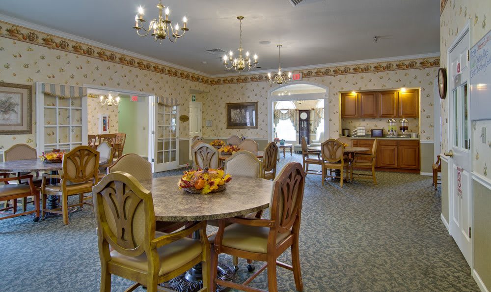 A cozy dining room with elegant chandeliers and wooden furniture, featuring a kitchen area in the background.