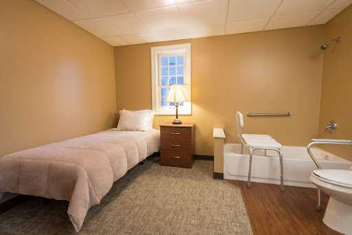 A cozy bedroom featuring a single bed, a nightstand with a lamp, and a chair, all set against warm-colored walls.