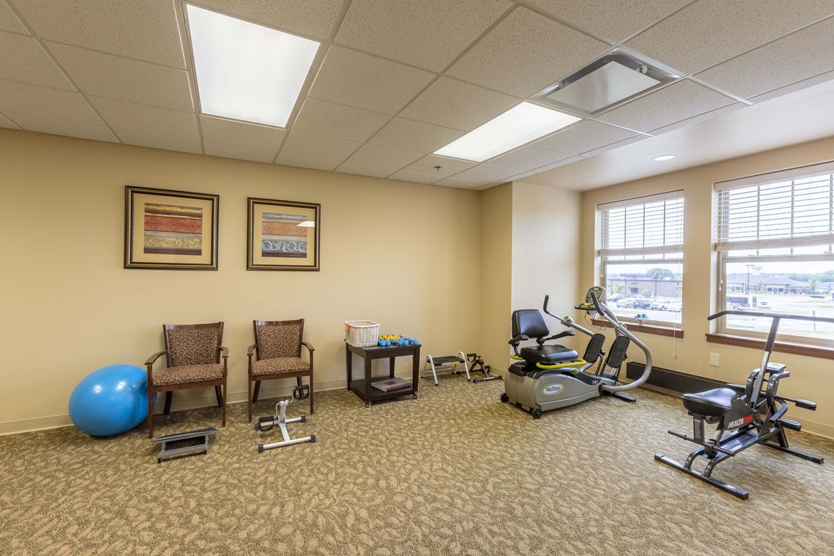 A small exercise room featuring a treadmill and stationary bike, with chairs and bright overhead lighting.