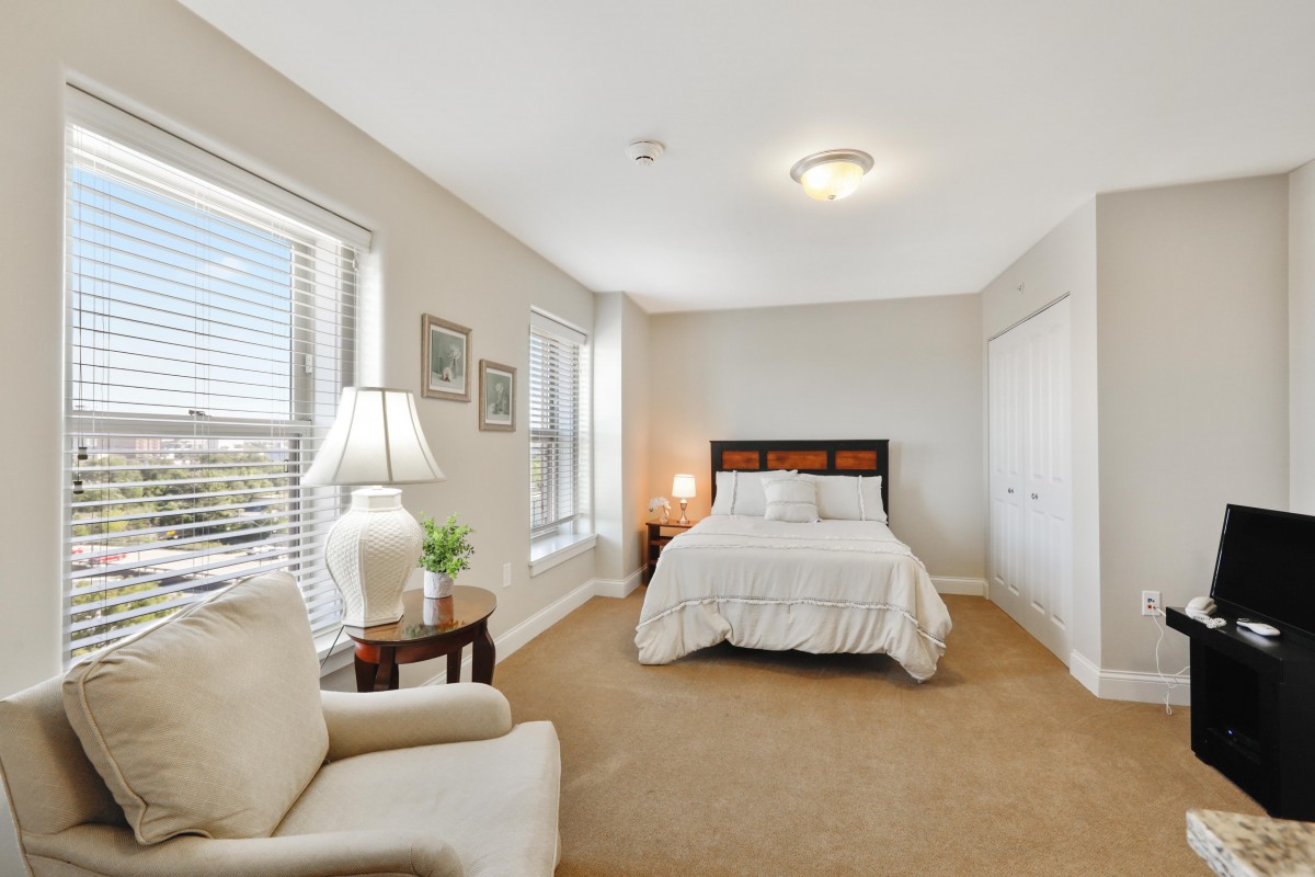 A cozy bedroom with a comfortable bed, an armchair, and ample natural light from large windows.