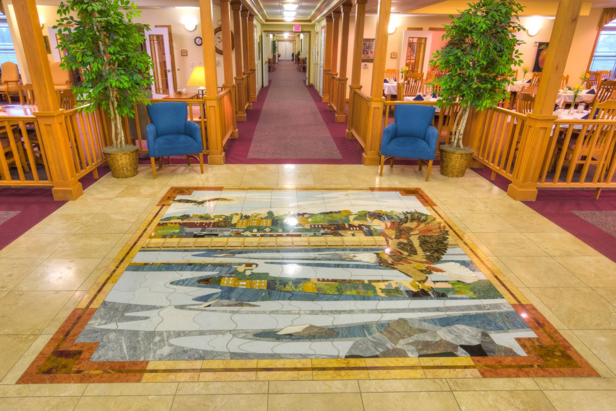 A hotel lobby featuring a decorative floor artwork, flanked by potted plants and blue chairs along the hallway.