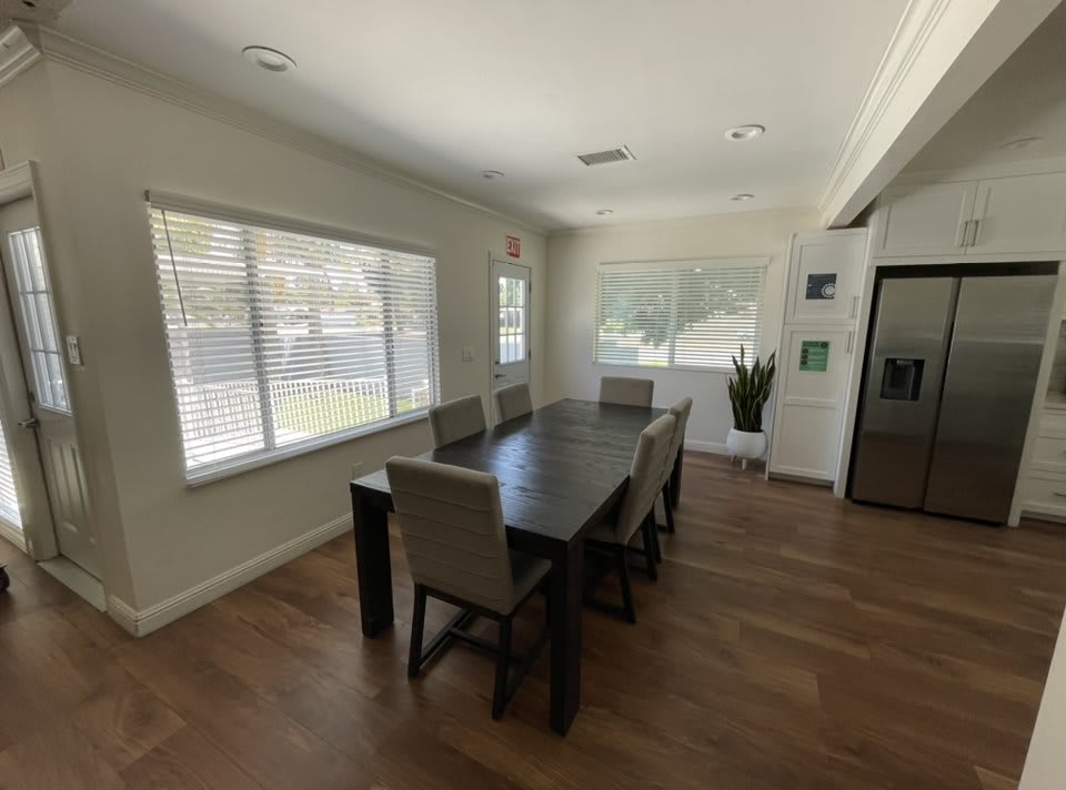 A bright dining room with a dark wooden table and six chairs, featuring large windows and wood flooring.