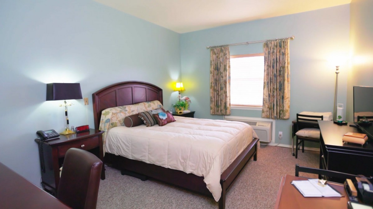 A cozy bedroom with a large bed, wooden headboard, and soft pillows. A nightstand with a lamp is beside the bed, and curtains cover the window, creating a warm atmosphere.