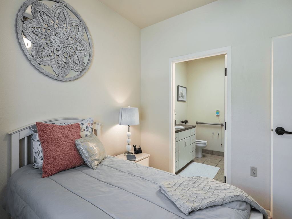 A cozy bedroom with a neatly made bed, decorative pillows, and a lamp on the bedside table. A doorway leads to an adjoining bathroom.