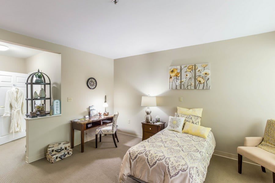 A cozy bedroom featuring a single bed, a desk with a chair, decorative artwork, and a bedside lamp.
