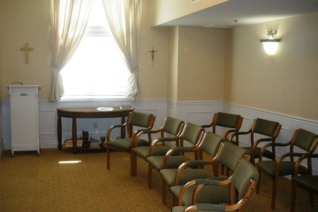 A small chapel with several chairs arranged in rows, a window with curtains, and religious symbols on the walls.