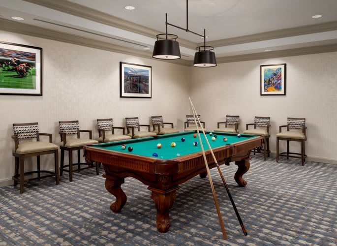 A well-lit game room featuring a billiard table surrounded by chairs, with cue sticks resting against it.