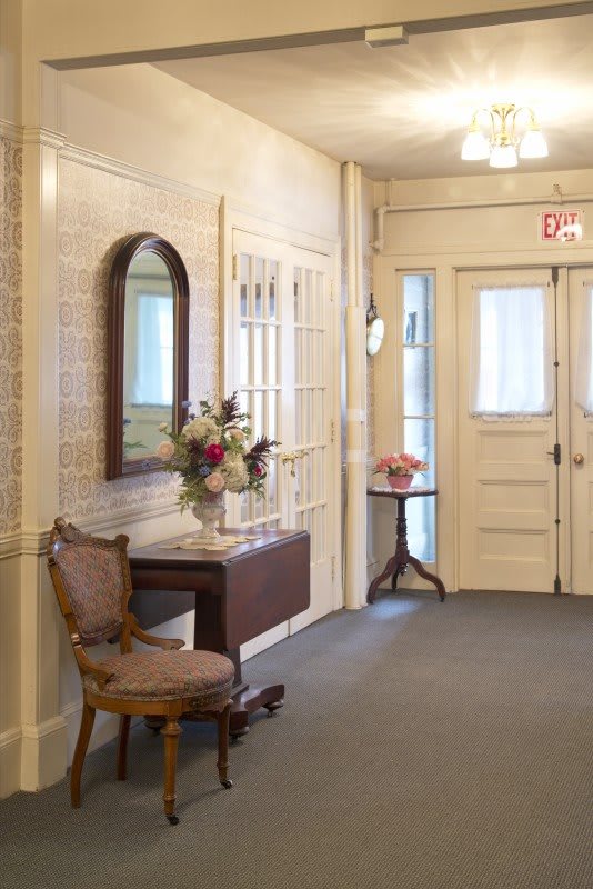 A vintage-style hallway featuring a chair, a small table with flowers, and a mirror.