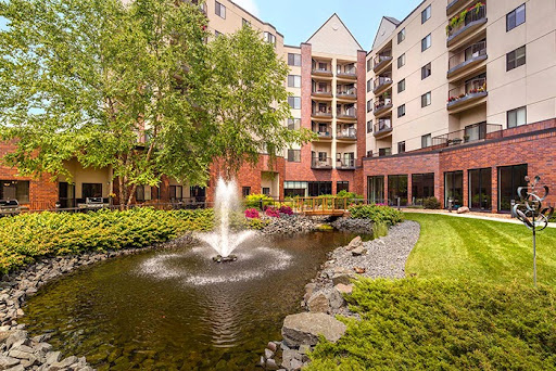 A serene view of a landscaped pond featuring a water fountain, surrounded by an apartment complex.