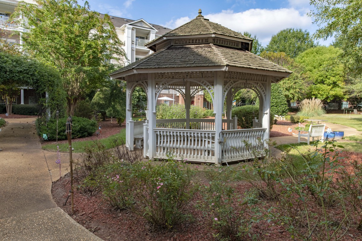 A charming wooden gazebo surrounded by a well-maintained garden area, set against a backdrop of trees and residential buildings.