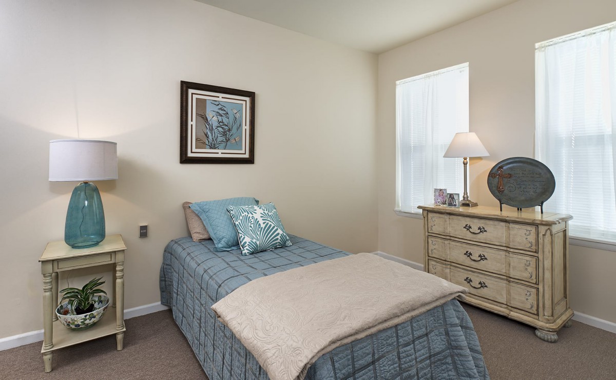 A cozy bedroom with a single bed, featuring decorative pillows and a neatly arranged dresser with a lamp.