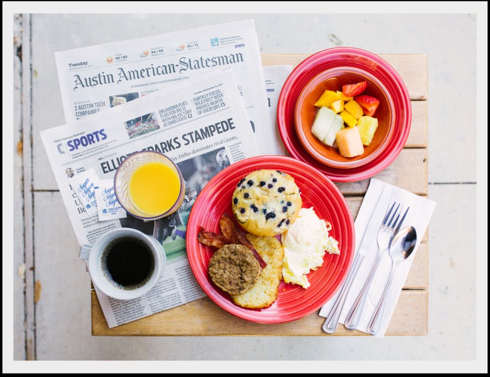 A breakfast spread with a newspaper, featuring sausage, potatoes, eggs, a muffin, fruit salad, coffee, and orange juice.