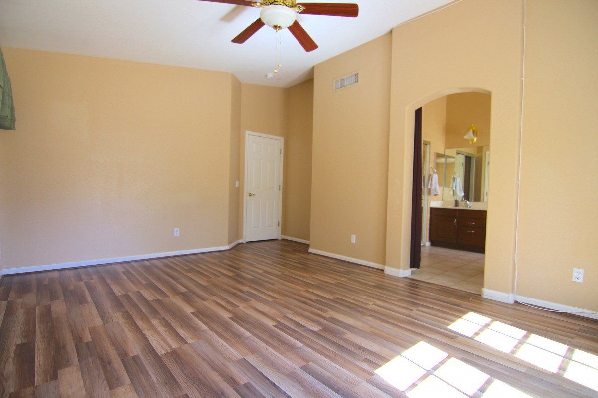 An empty room with hardwood floors and a ceiling fan, featuring beige walls and a doorway leading to another area.
