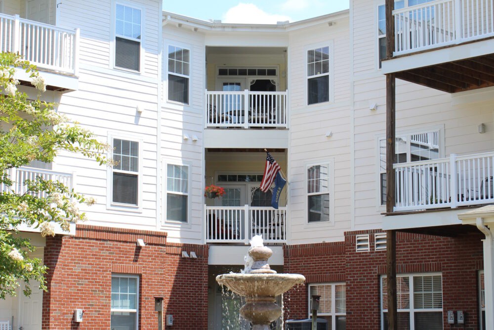 A residential apartment building with multiple balconies and an American flag displayed, featuring a fountain in the foreground.