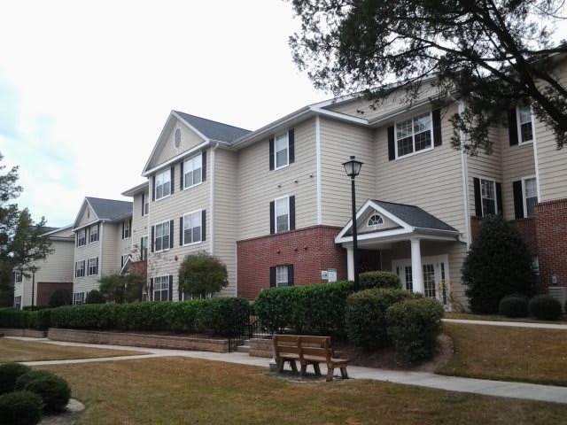 A suburban apartment building with surrounding greenery and a bench in the foreground.
