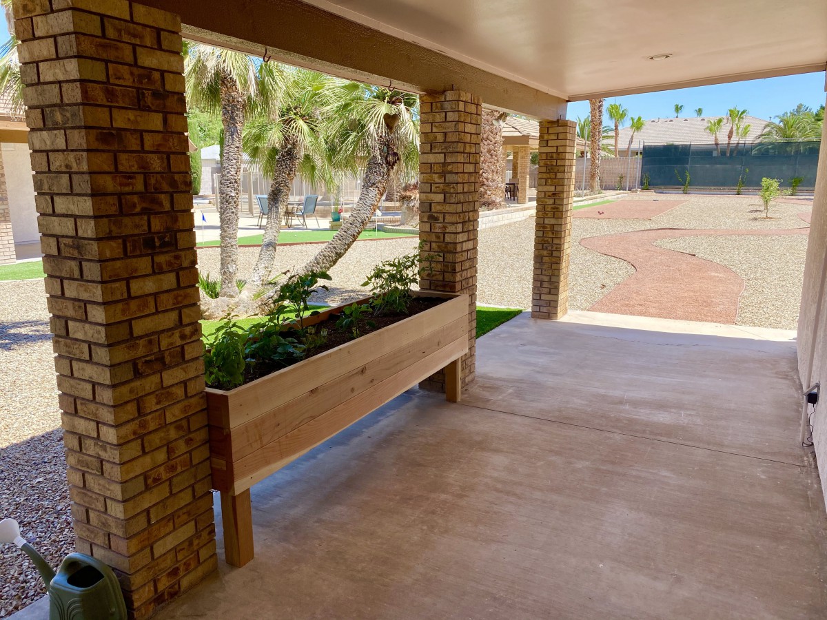 A wooden planter box filled with greenery on a covered patio with brick columns.