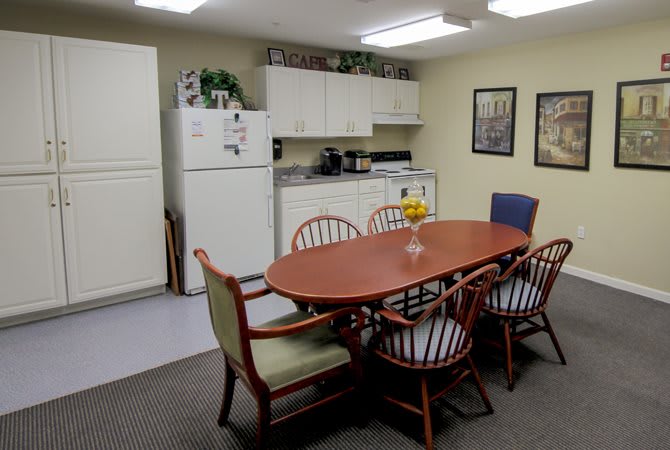 A small office kitchen with a dining table and chairs, featuring a refrigerator and overhead cabinets.