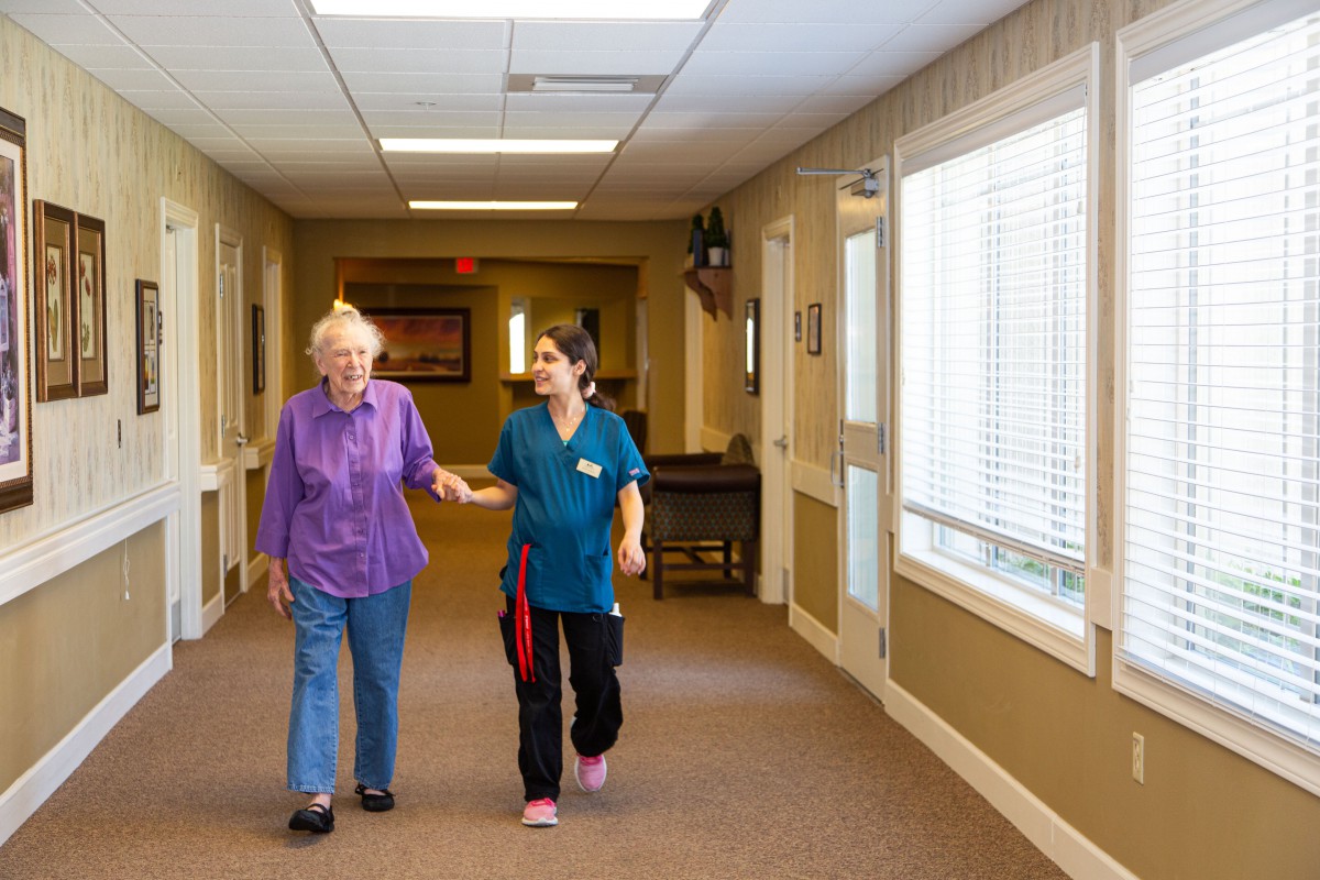 A caregiver assisting an elderly person in a nursing home hallway.