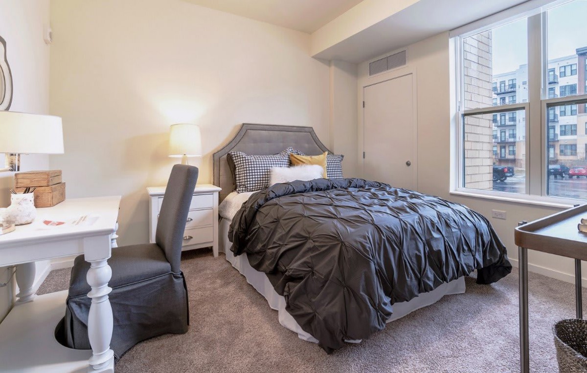 A cozy bedroom featuring a neatly made bed with dark bedding, a desk with a chair, and a lamp providing light.