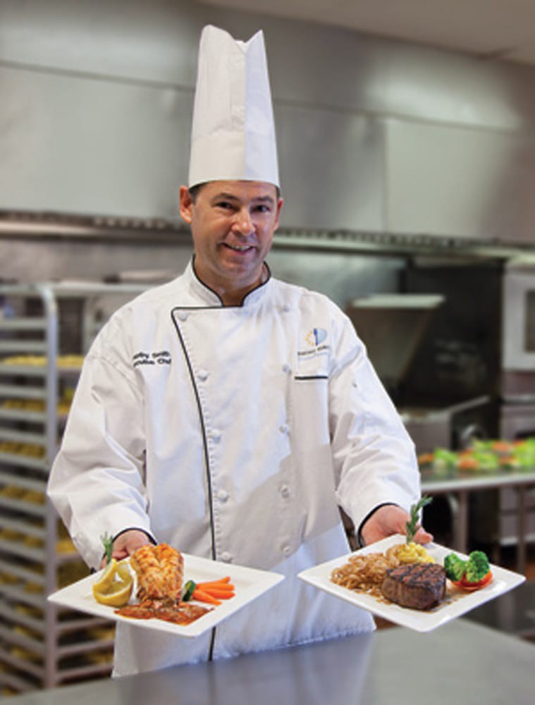 A chef presents two elegantly plated dishes in a professional kitchen.