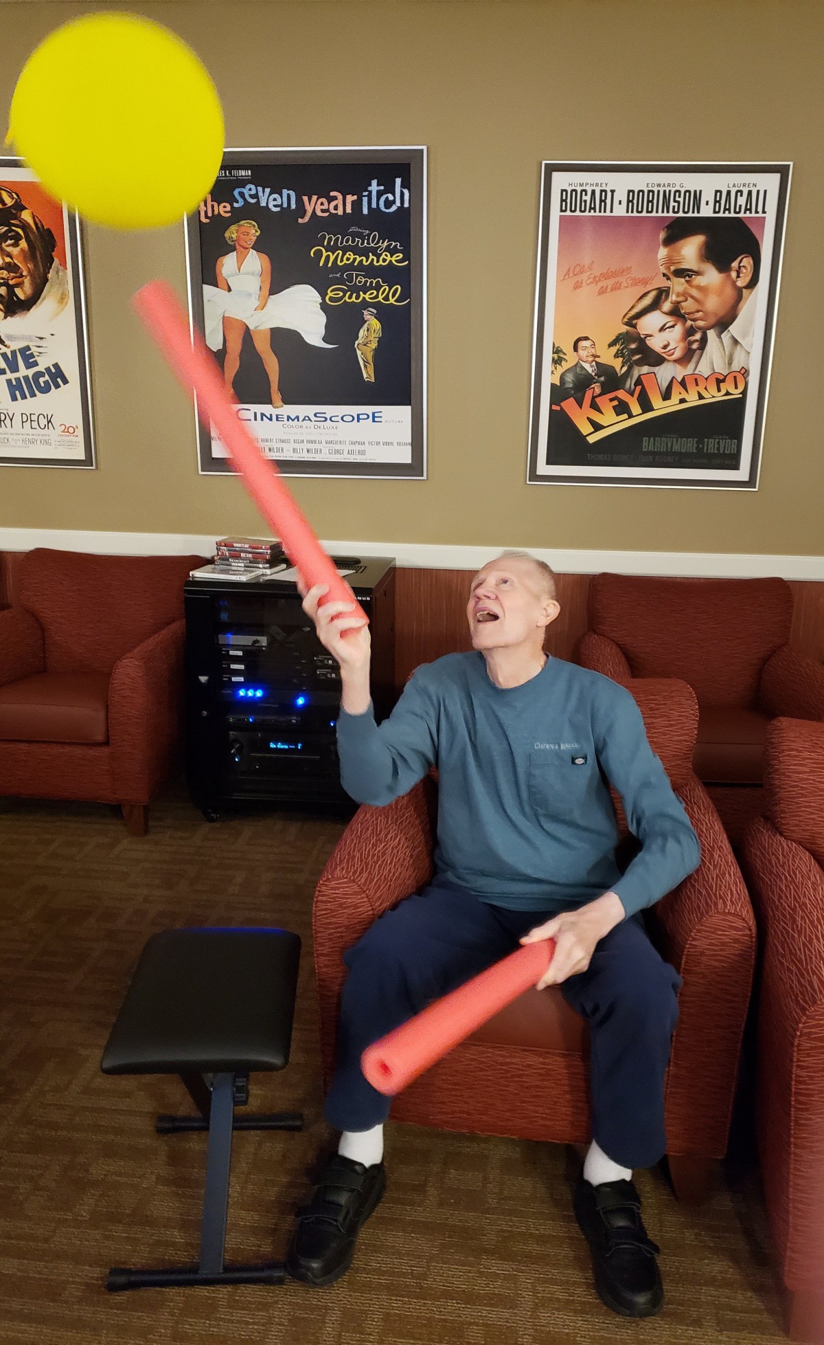 A person sitting on a chair indoors, playfully holding two foam noodles with movie posters in the background.