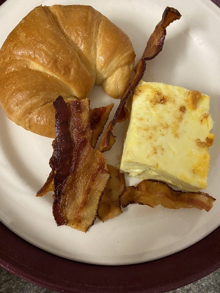 A breakfast plate featuring crispy bacon, a croissant, and a square of egg casserole.