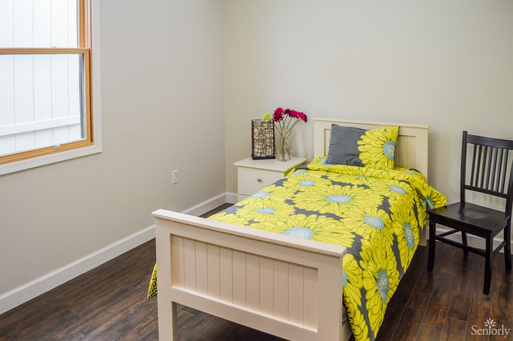 A small bedroom featuring a single bed with bright yellow bedding, complemented by a decorative pillow and a side table with a lamp and flower vase.