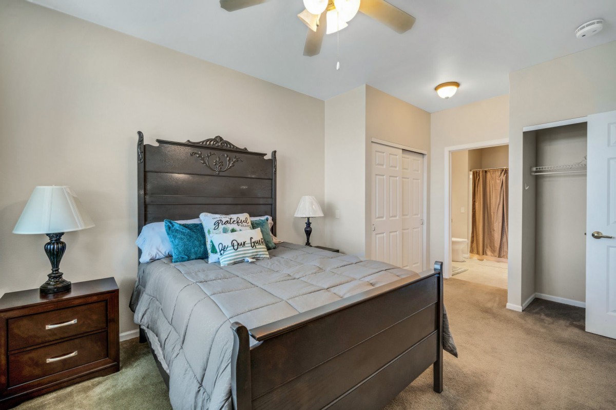 A cozy bedroom featuring a wooden bed with decorative pillows, a bedside lamp, and a ceiling fan. An open door leads to an adjacent room.