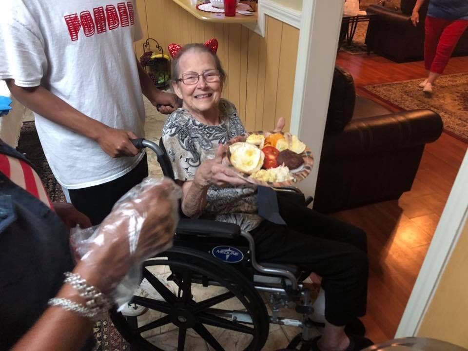An elderly woman in a wheelchair is holding a birthday cake and smiling during a celebration.