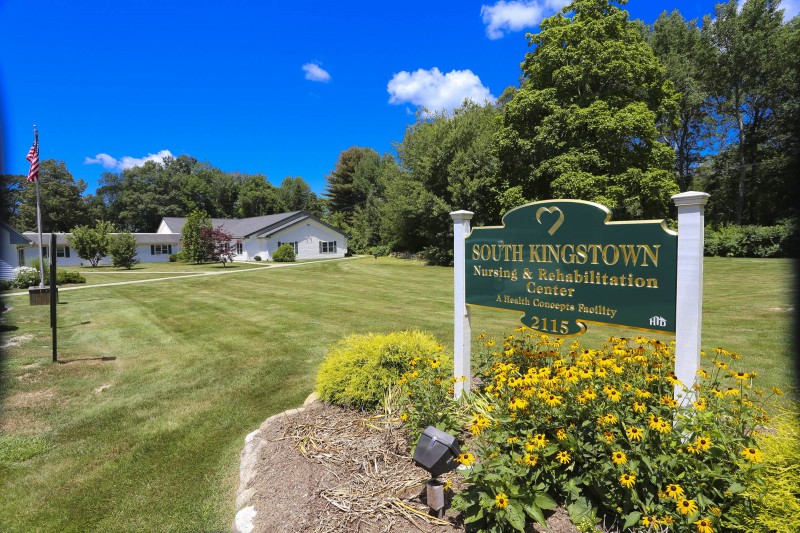 View of a sign and building surrounded by greenery and flowers under a clear blue sky.