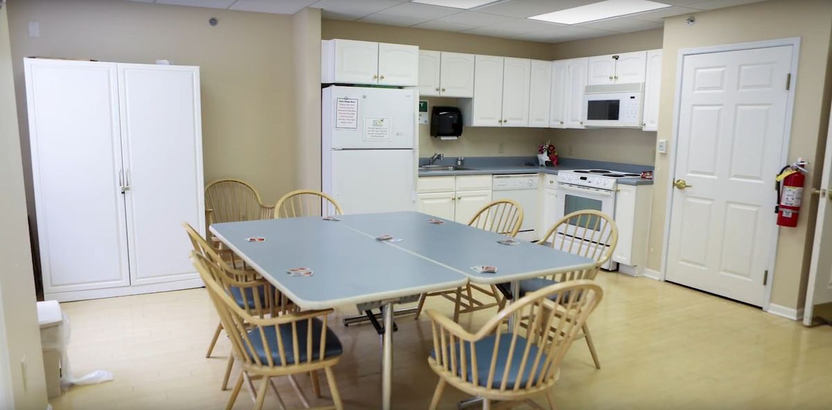 A communal kitchen area with a large dining table and wooden chairs, surrounded by white cabinets and appliances.