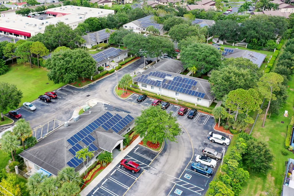 Aerial view of a complex with solar panels installed on rooftops and parking lot canopies, surrounded by greenery.