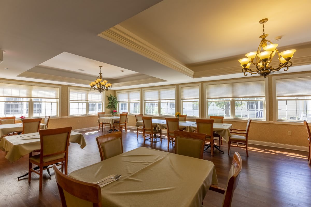 A well-lit dining area with neatly arranged tables and chairs, featuring large windows and elegant chandeliers.