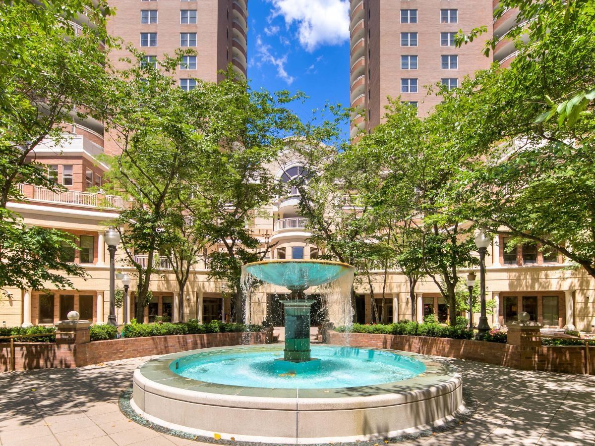 A scenic courtyard with a central fountain surrounded by trees and tall buildings.
