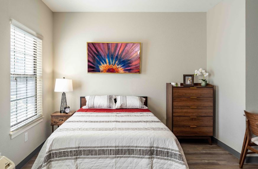 A cozy bedroom with a neatly made bed, featuring a colorful artwork above the headboard, a bedside lamp, and a wooden dresser.