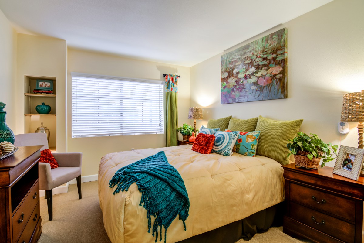 A cozy bedroom with colorful decor, featuring a large bed adorned with a variety of pillows and a vibrant blue throw blanket. The walls are decorated with a large piece of artwork above the headboard, and natural light streams in through a window.