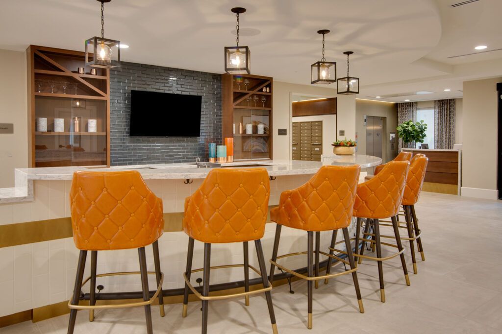 A modern kitchen area featuring a sleek countertop with vibrant orange bar stools, pendant lighting, and a wall-mounted television.