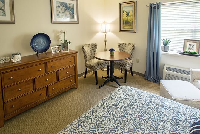 A cozy bedroom with a dresser, round table, and two chairs, adorned with decorative artwork and a floor lamp.