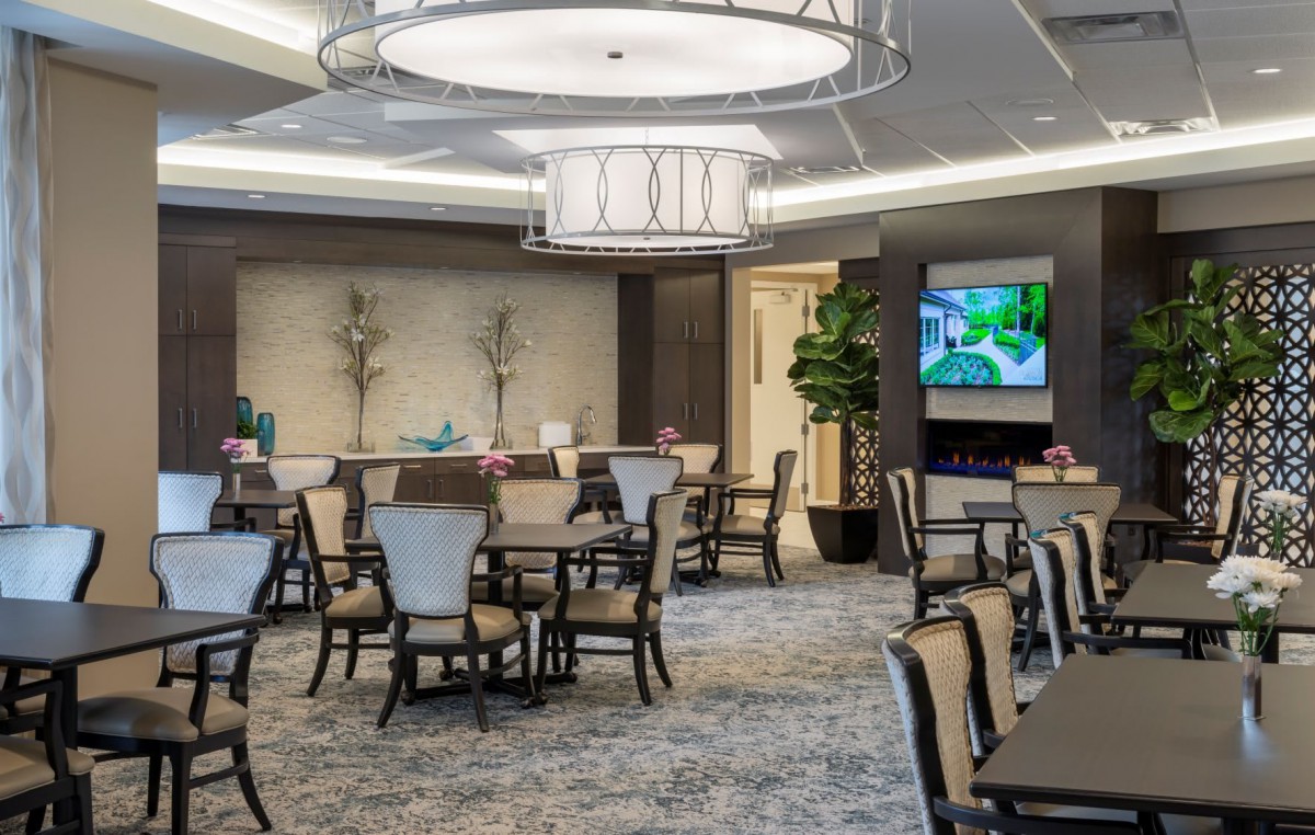 A well-lit dining room with elegant furniture and chandeliers, featuring tables and chairs arranged for dining.