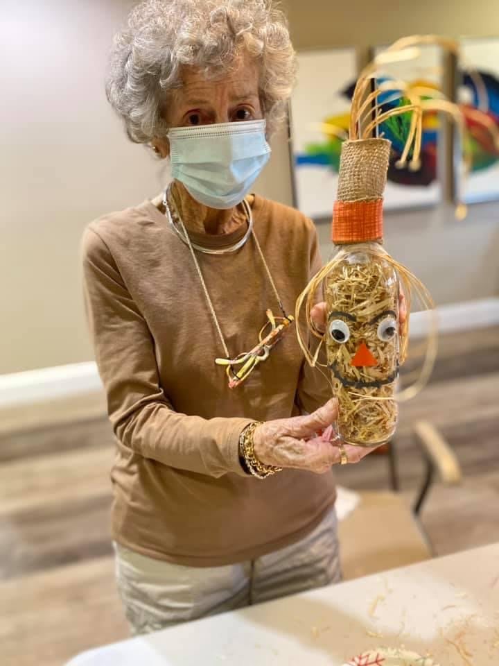 An elderly woman indoors, wearing a mask, holds up a handcrafted art piece.