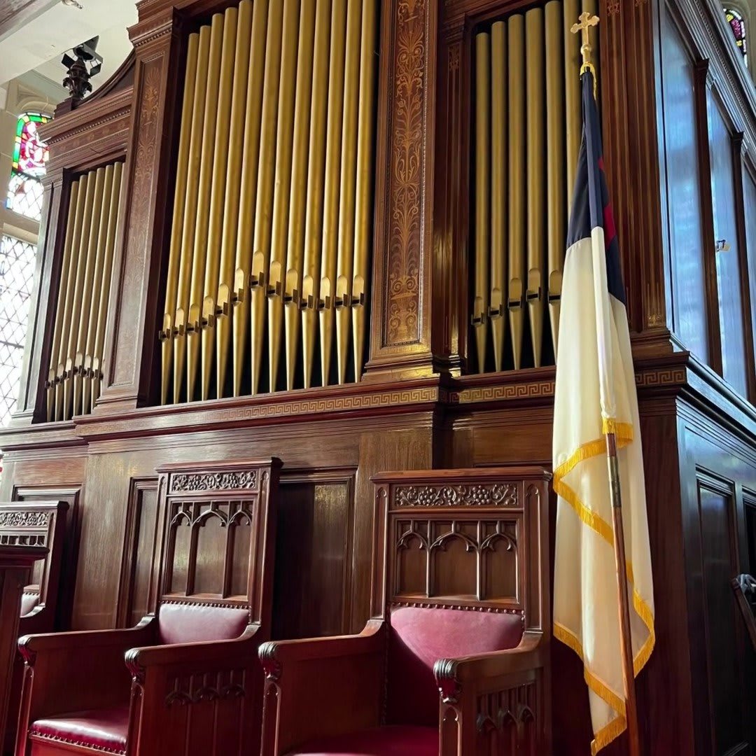 A church interior featuring a large organ with wooden panels and adjacent pews, accompanied by a flag.