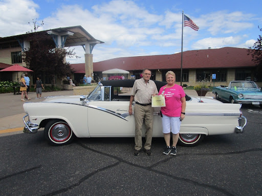 A couple celebrates their classic car at an outdoor event, proudly displaying a certificate next to their vintage vehicle.