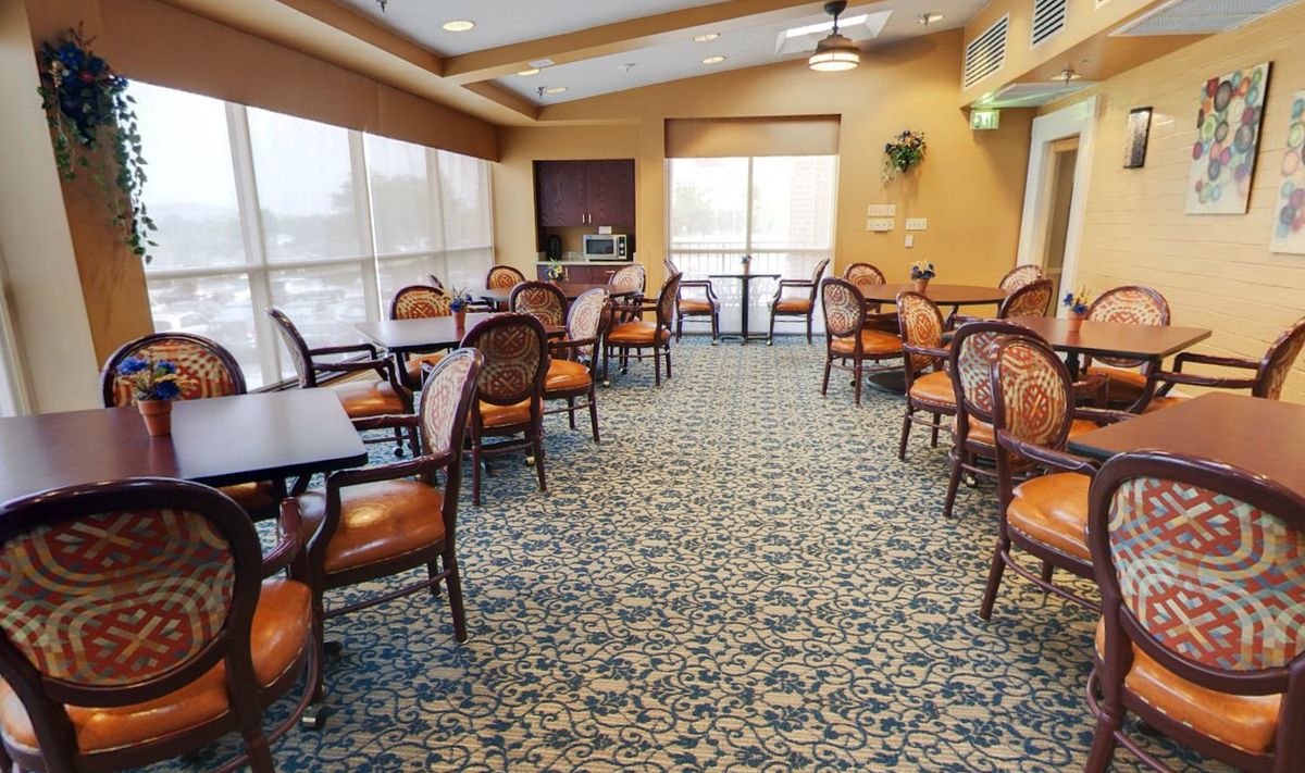 A spacious and elegantly decorated dining area featuring multiple tables and chairs, with carpeted flooring and large windows allowing natural light.