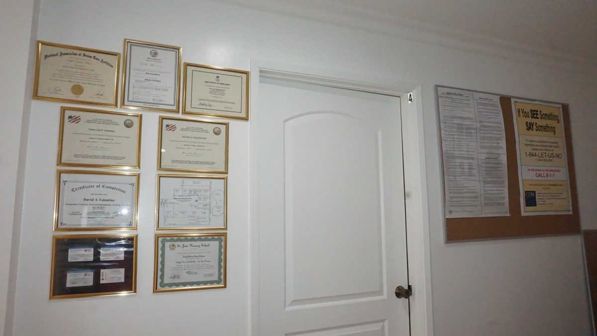 A collection of framed certificates and diplomas displayed on a white wall next to a closed door in an office setting.