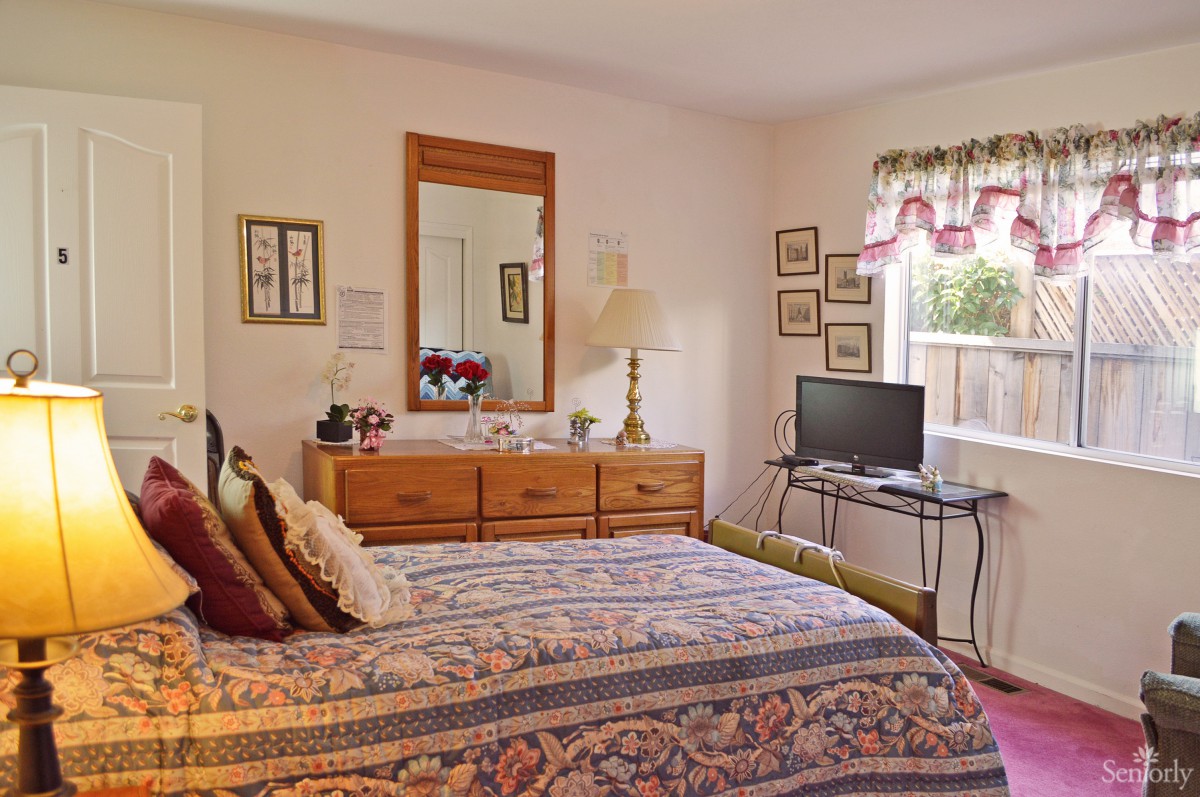 A cozy bedroom with a bed, dresser, and TV, featuring floral decor and natural light.