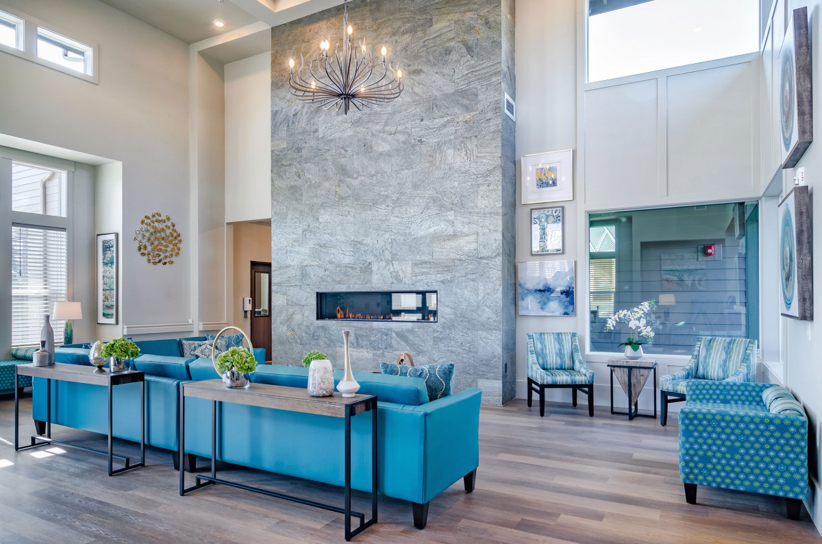 A spacious, modern living room with blue sofas and a central fireplace, beautifully lit by a chandelier and natural light from large windows.