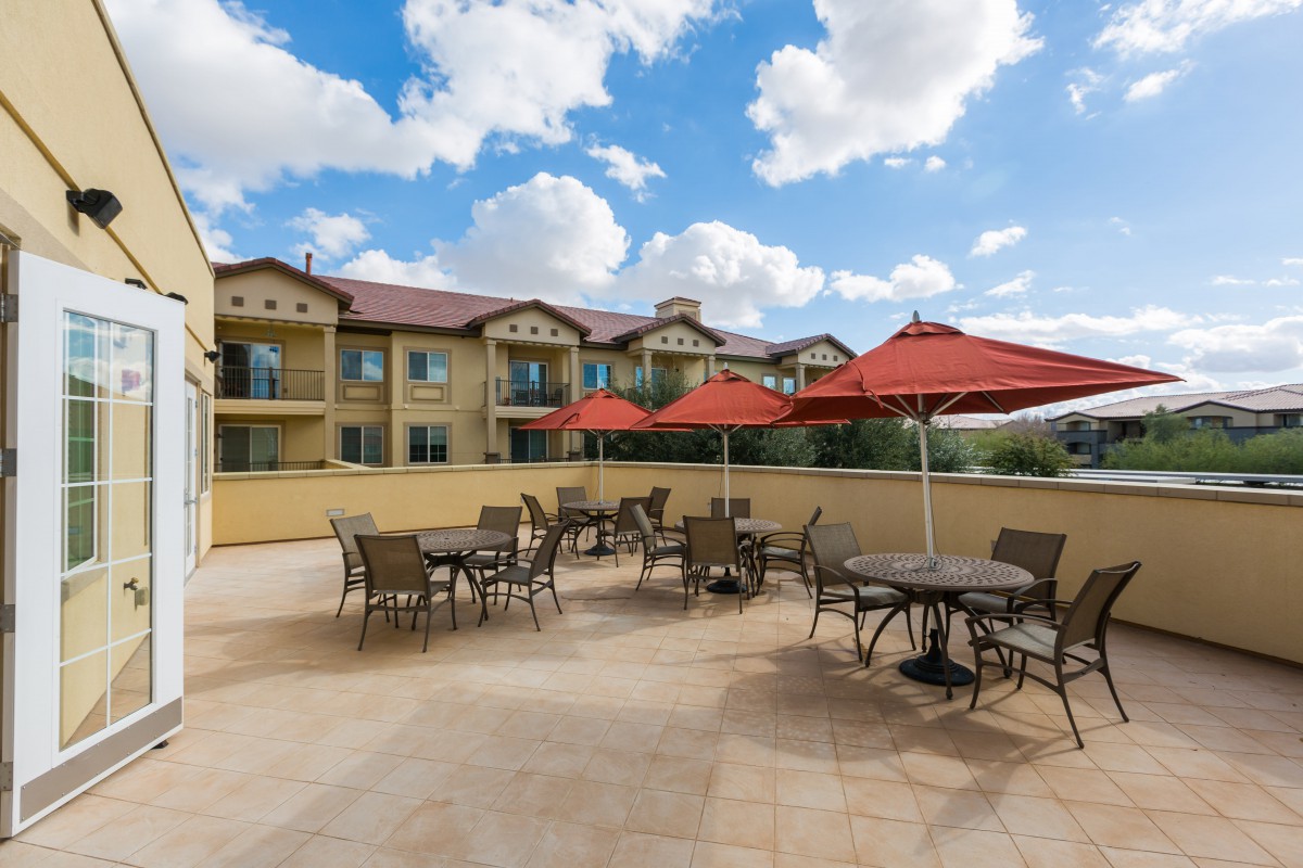 An outdoor patio with tables, chairs, and red umbrellas set against a building exterior under a partly cloudy sky.